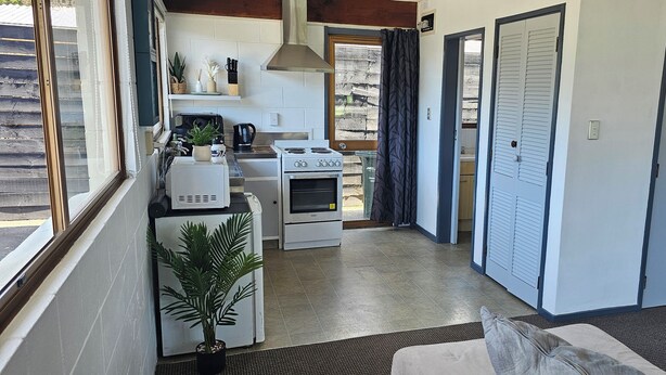 A compact kitchen with a stove, oven, and sink, a dining area with a table and chairs, and a small seating area with a sofa and cushions. - Cozy Cove Ohope Beach (Ohope)