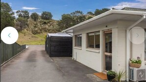 A small, single-story building with a white exterior, a covered porch, and a small garden area with a potted plant. - Cozy Cove Ohope Beach (Ohope)