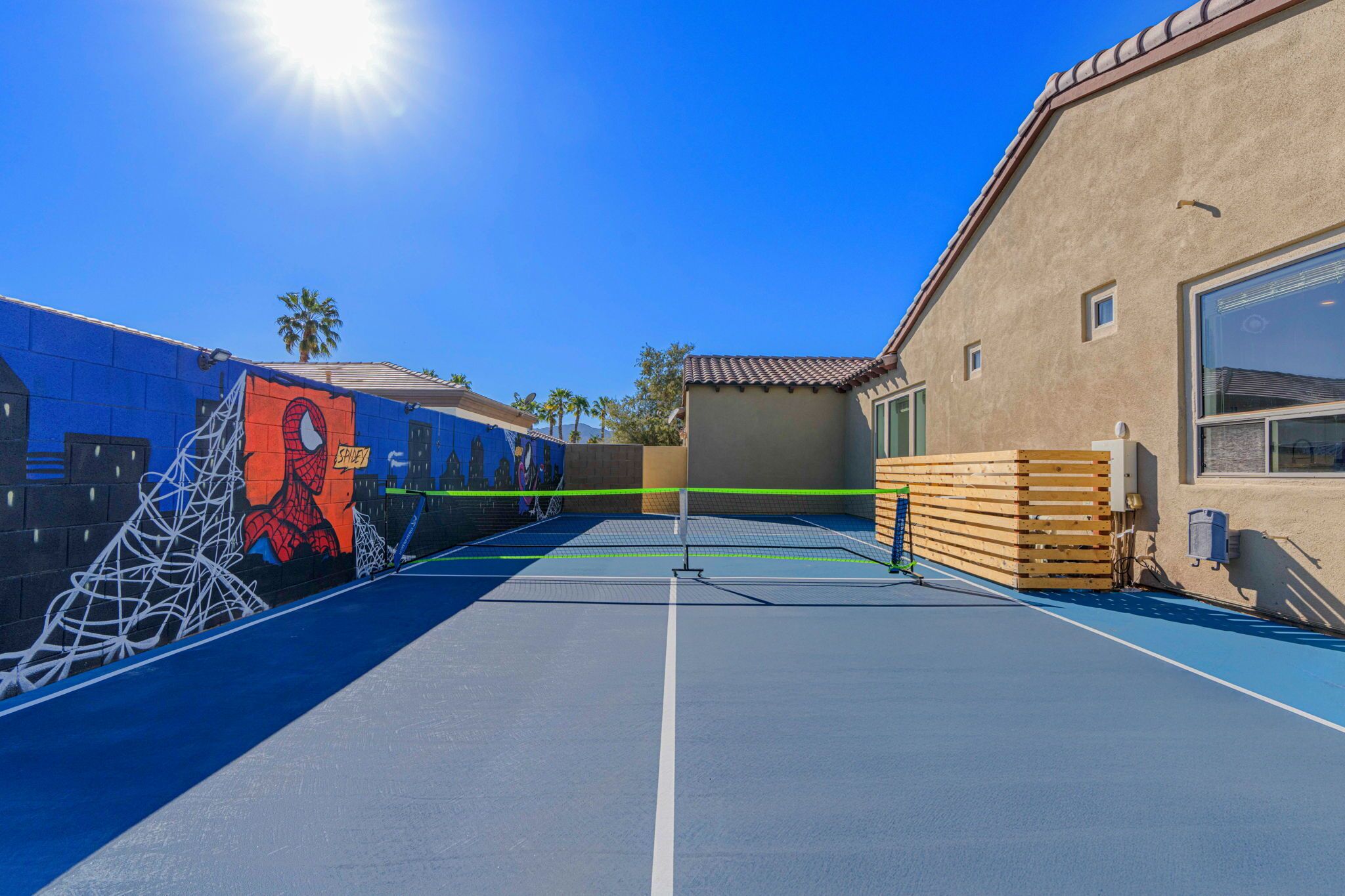 Un terrain de tennis en plein air, doté d’un filet, orné d’une fresque représentant Spider-Man, et un bâtiment avec un toit en tuiles.