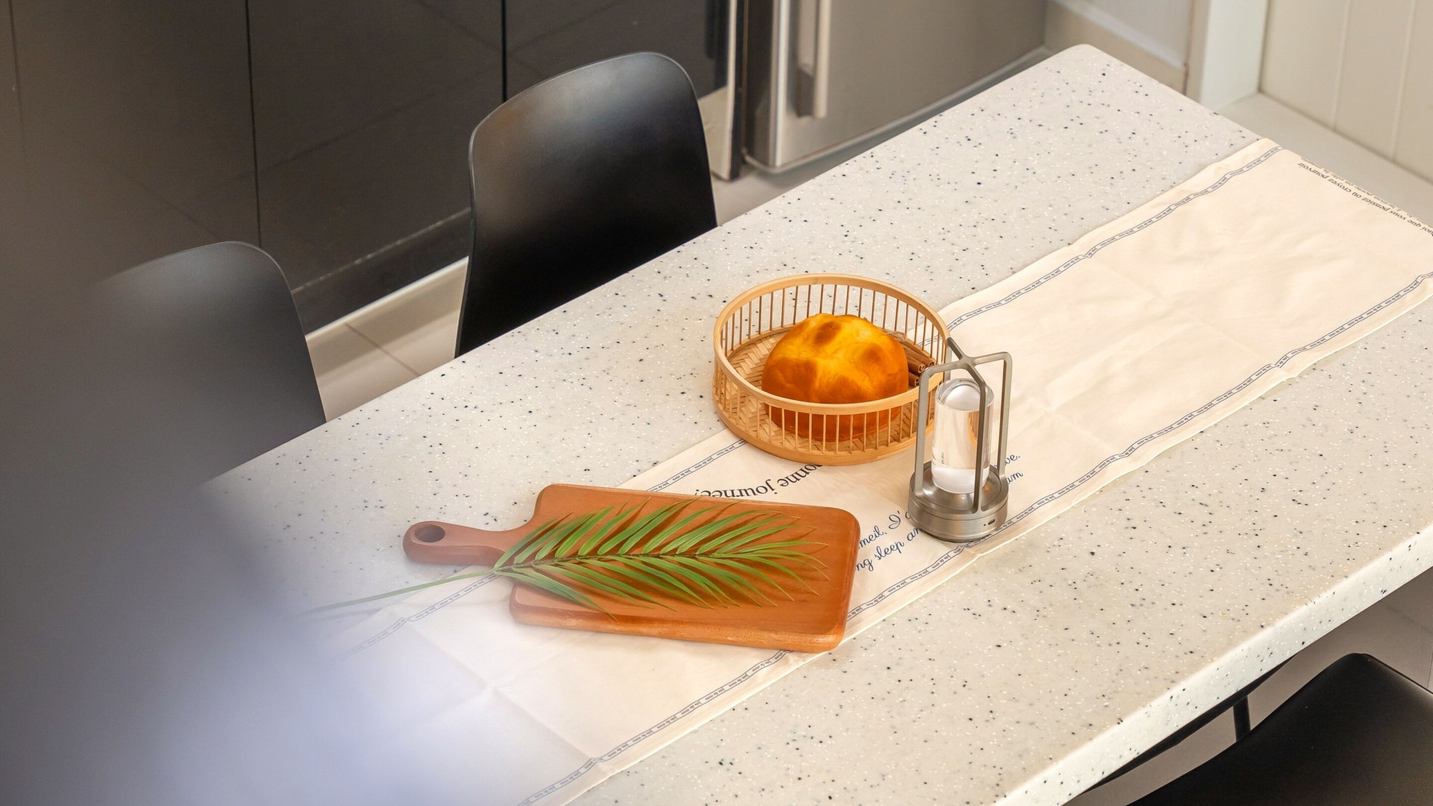 A kitchen counter with a white speckled backsplash, a wooden cutting board with green leaves, a woven basket with a loaf of bread, and a metal candle holder.