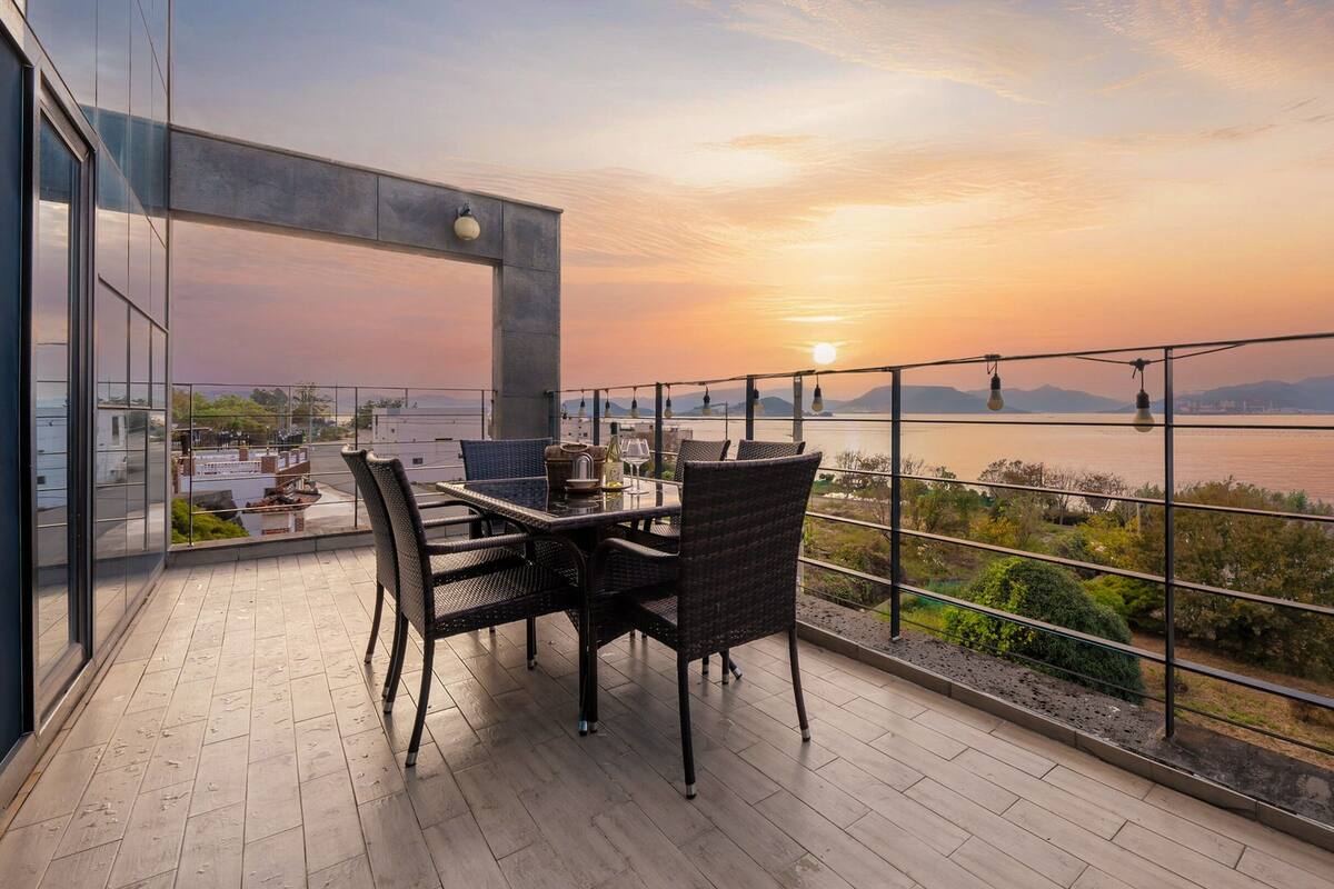 A balcony with a table and chairs, overlooking a body of water and distant mountains during sunset.