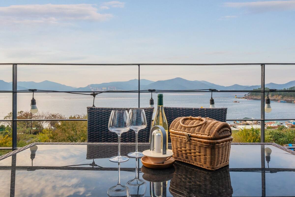 A table with two wine glasses, a bottle, and a basket, overlooking a body of water with boats and mountains in the distance.