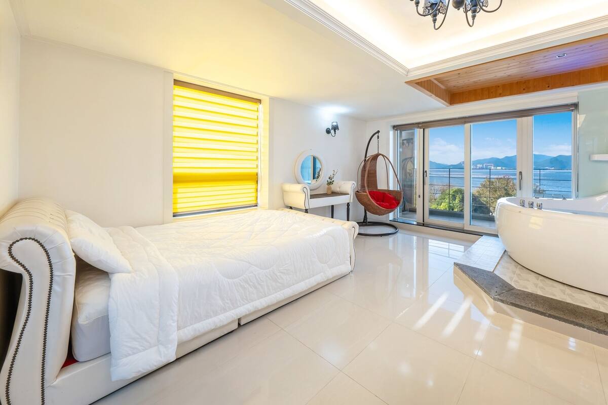 A modern bedroom with a bed, a hanging chair, a bathtub, and a view of the sea.