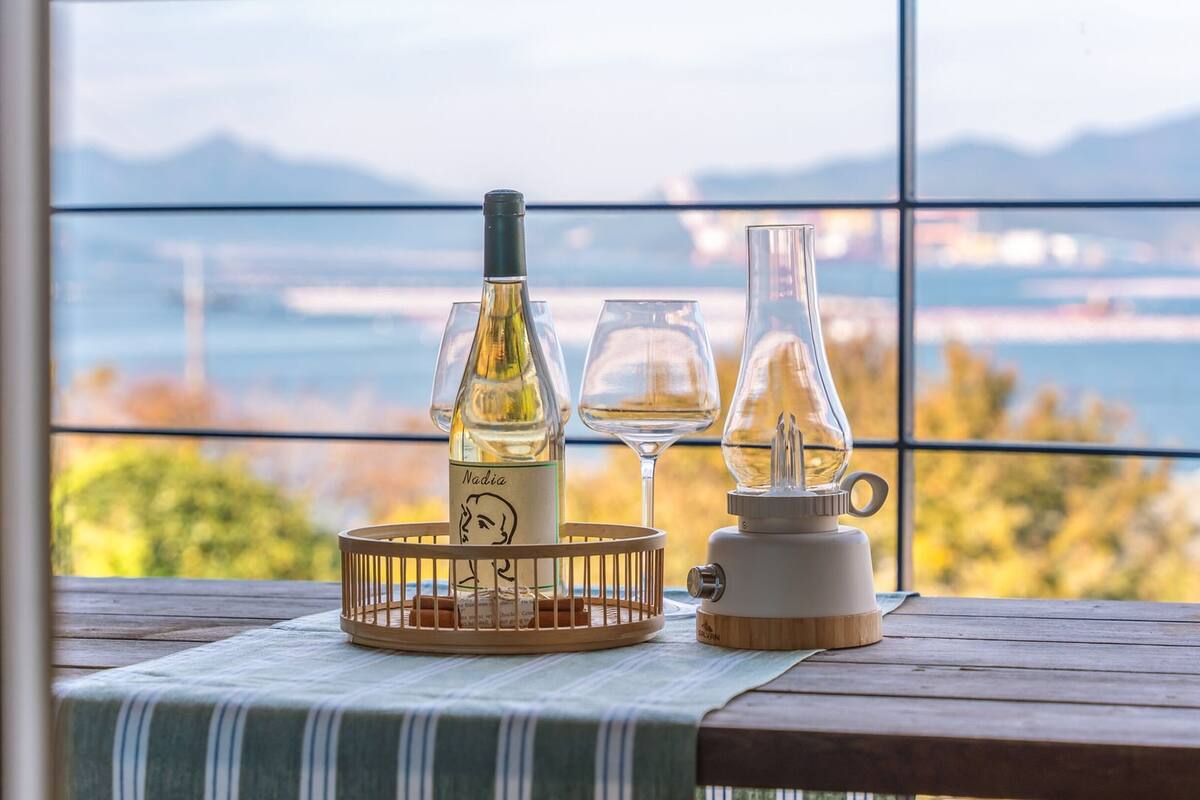 A table with a bottle of wine, a glass, and a candle holder by a window with a view of a body of water and mountains.