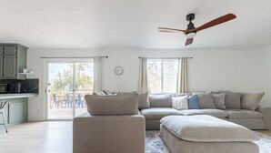A spacious living room with a large sectional sofa, a small ottoman, a ceiling fan, and a view of the outdoors through large windows.