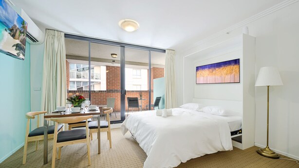 A hotel room with a bed, a desk with chairs, a table with a vase of flowers, and a painting on the wall. - MetaWise 2Bed Next to ICC Darling Harbour 65 (Sydney)