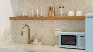 Una cocina con un lavamanos blanco, un grifo dorado, un microondas y una repisa con cristalería y una cafetera.