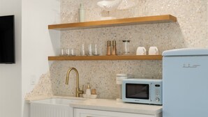 Una cocina con un lavamanos blanco, un grifo dorado, un microondas y un refrigerador. Hay estantes de madera sobre el lavamanos que contienen diversos utensilios de cocina.