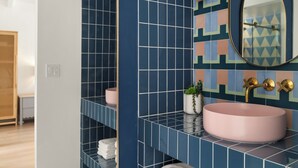 Un baño con un lavamanos redondo rosa, azulejos azules y multicolores, un espejo y una pequeña planta en maceta.