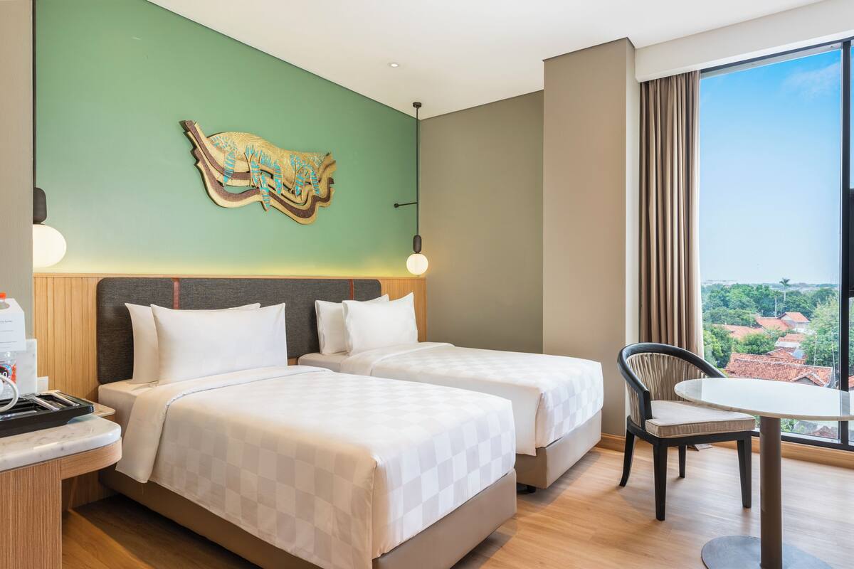 Ein Hotelzimmer mit zwei Betten, einem Esstisch, einem Sessel, einem großen Fenster mit Ausblick und einem Wandbild.
