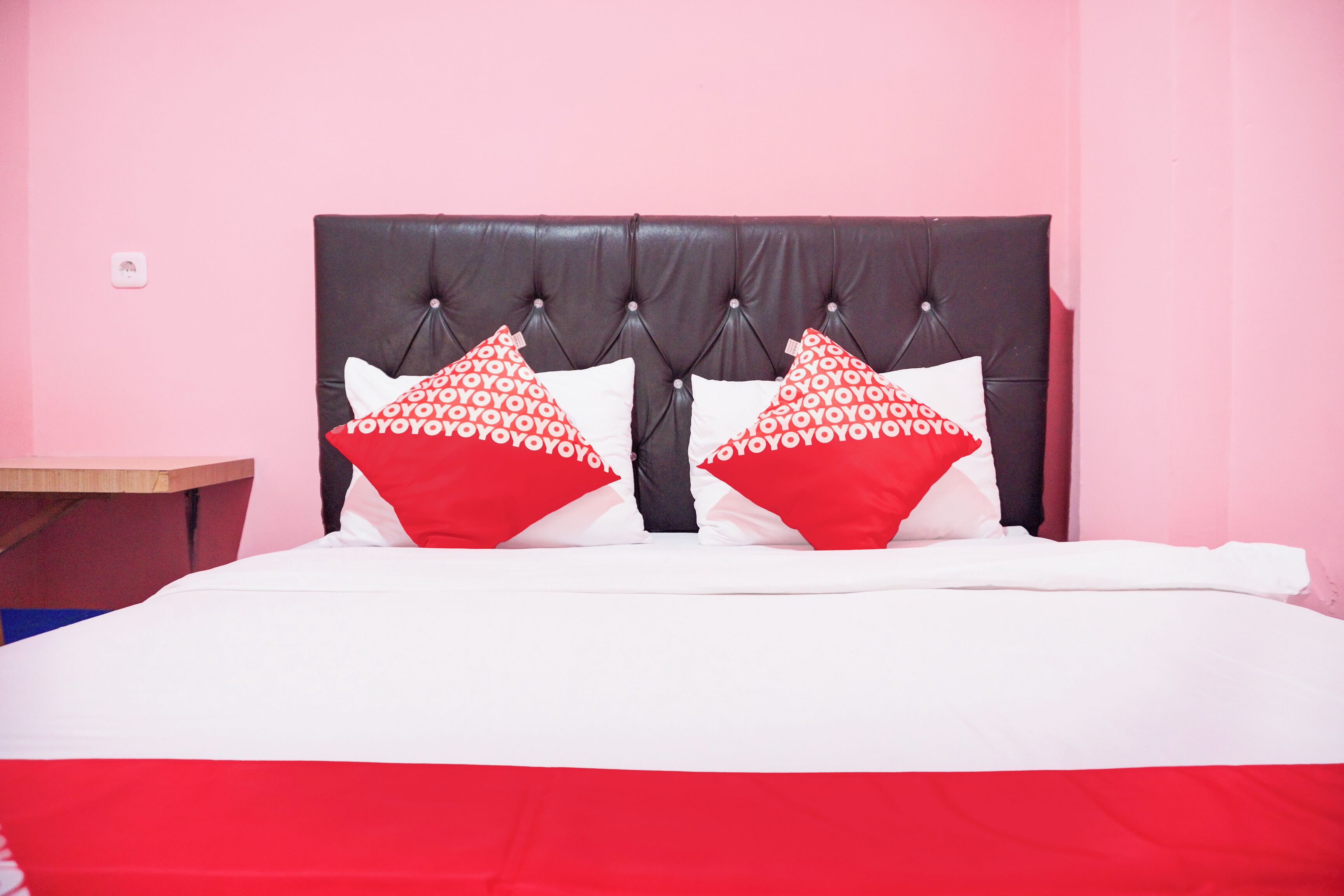 Una cama con cabecera capitonada, dos almohadas rojas con diseño blanco y una colcha blanca.