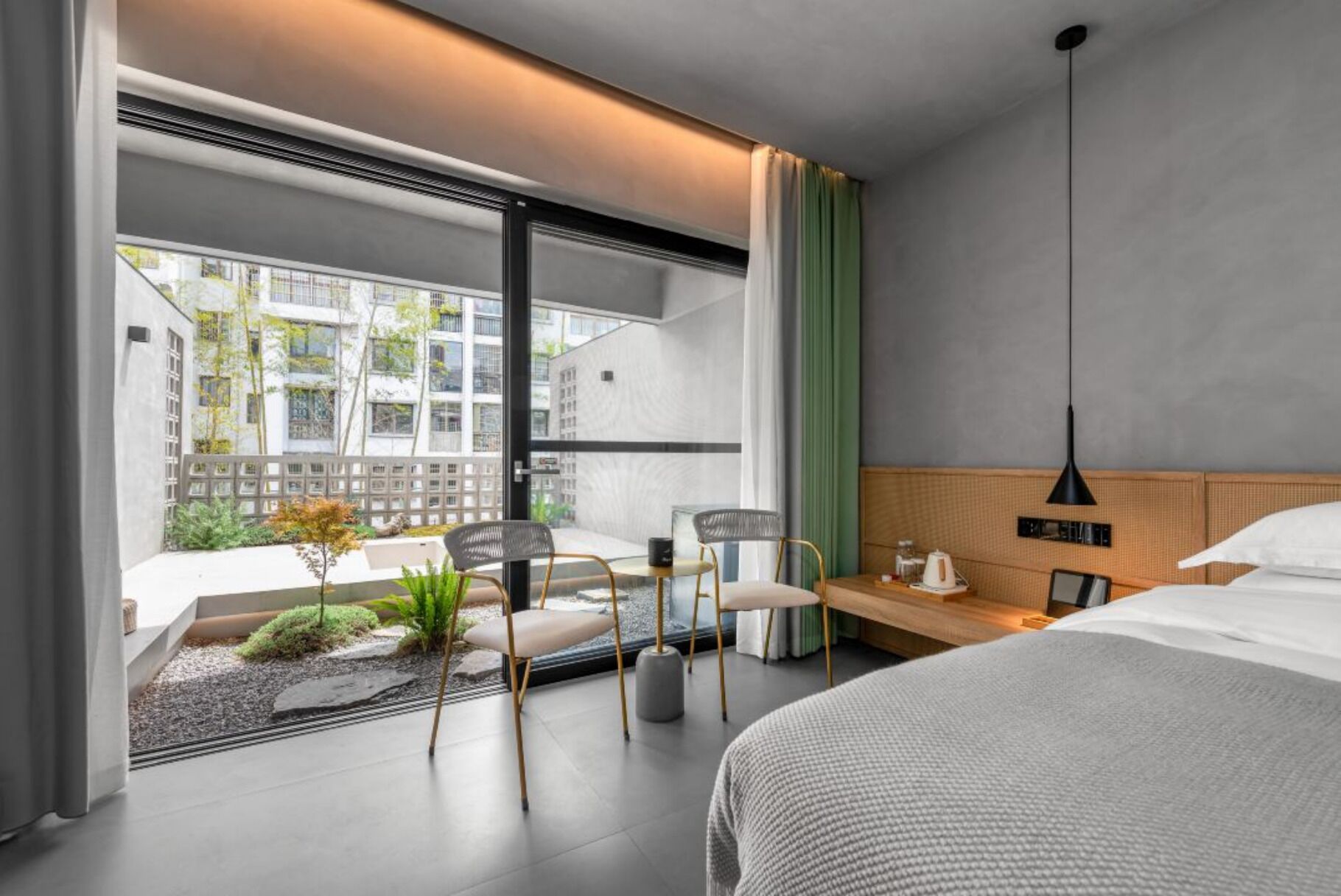 A modern hotel room with a bed, a desk with a chair, a view of the city, and a balcony with plants.