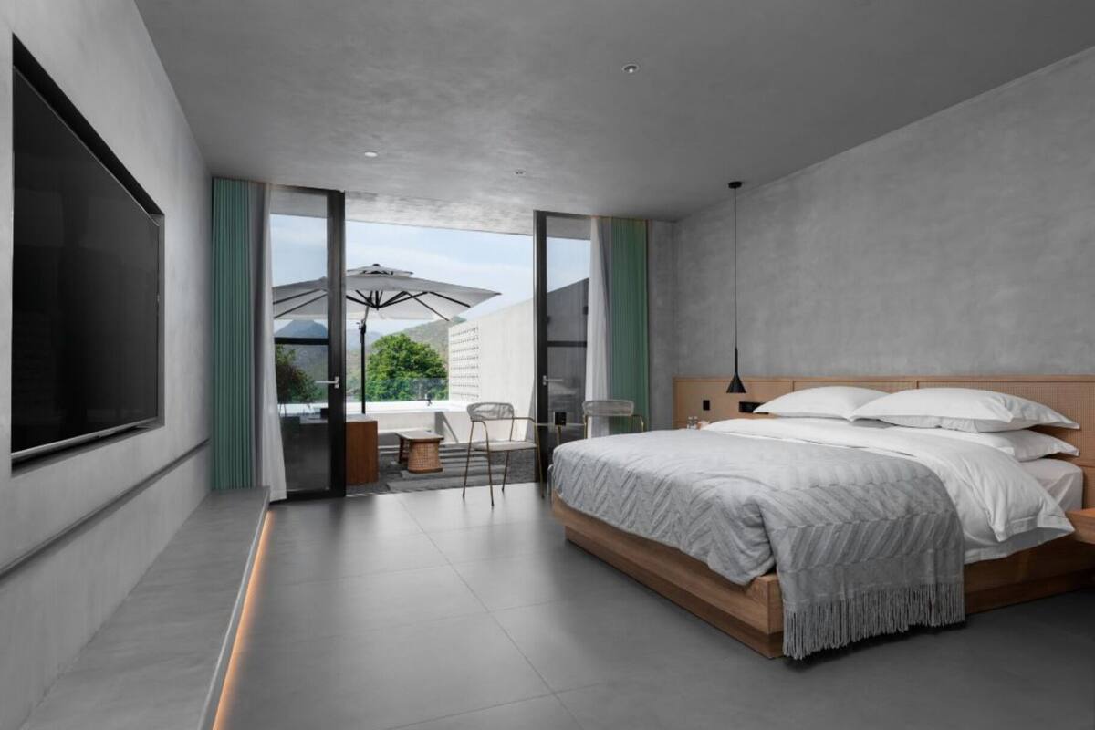 Une chambre moderne avec un grand lit, un balcon avec un fauteuil et une vue sur l’extérieur.