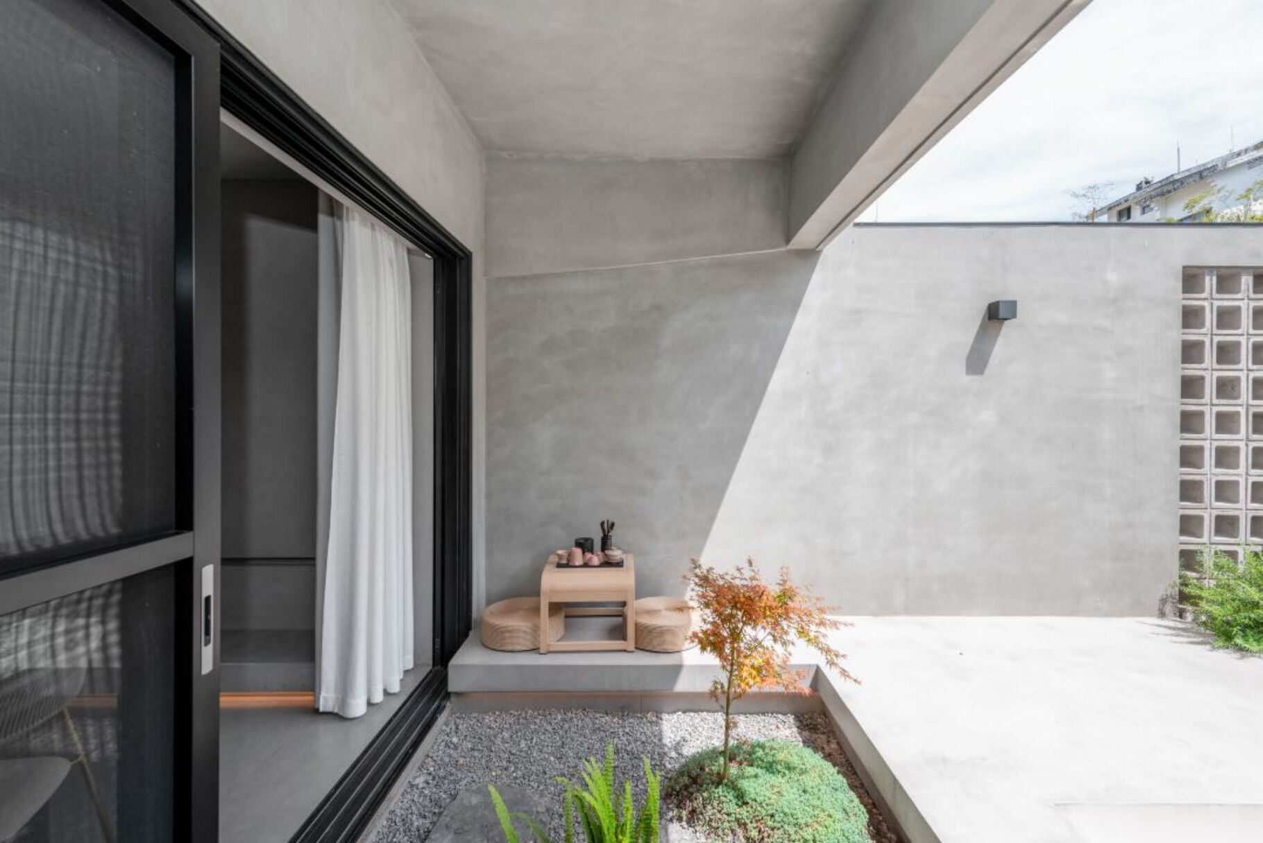 A modern, minimalist outdoor space with a concrete wall, a small seating area with cushions, a wooden stool, and a small tree.