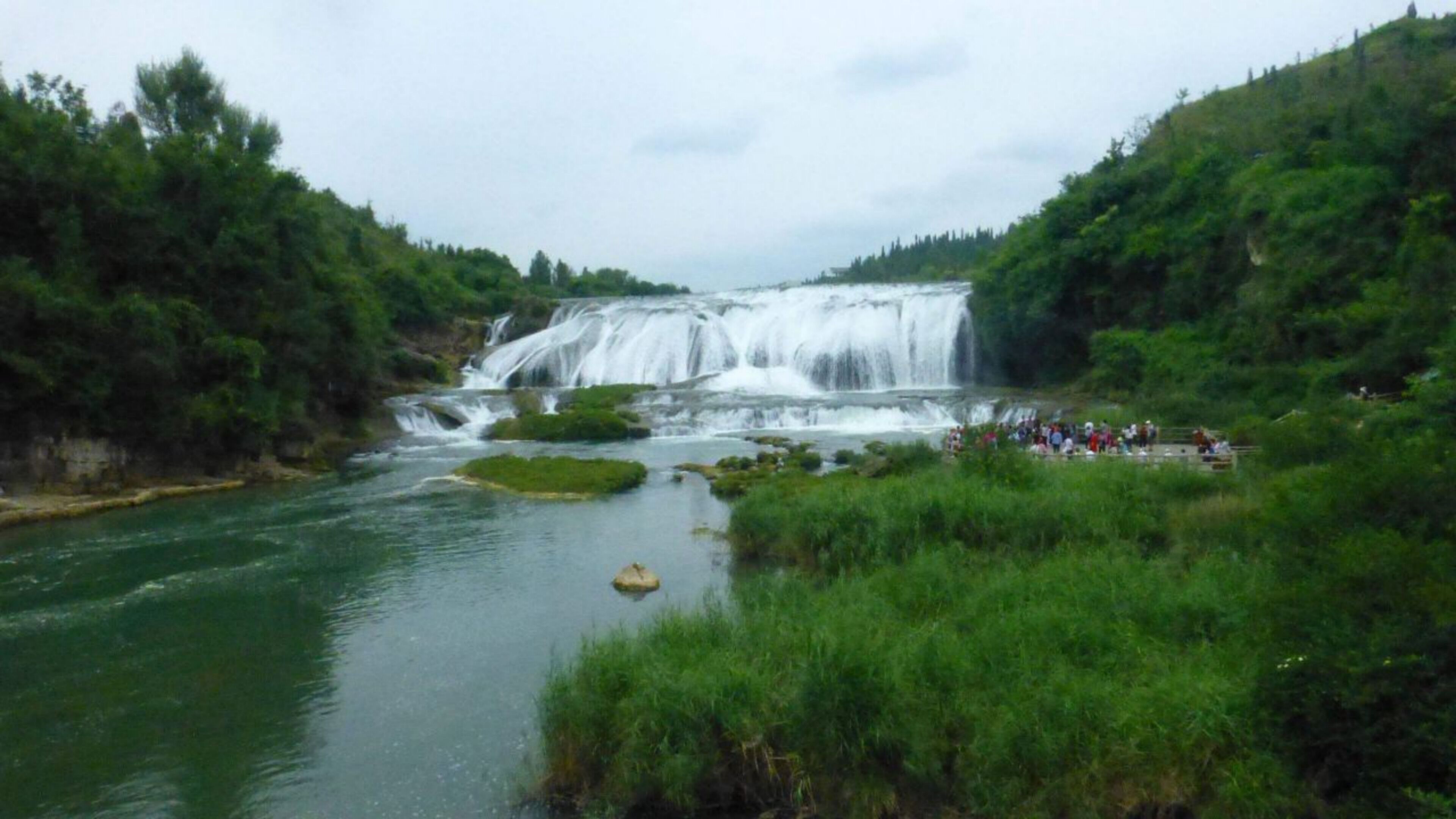 A waterfall with a river in the foreground and a crowd of people gathered at the base.
