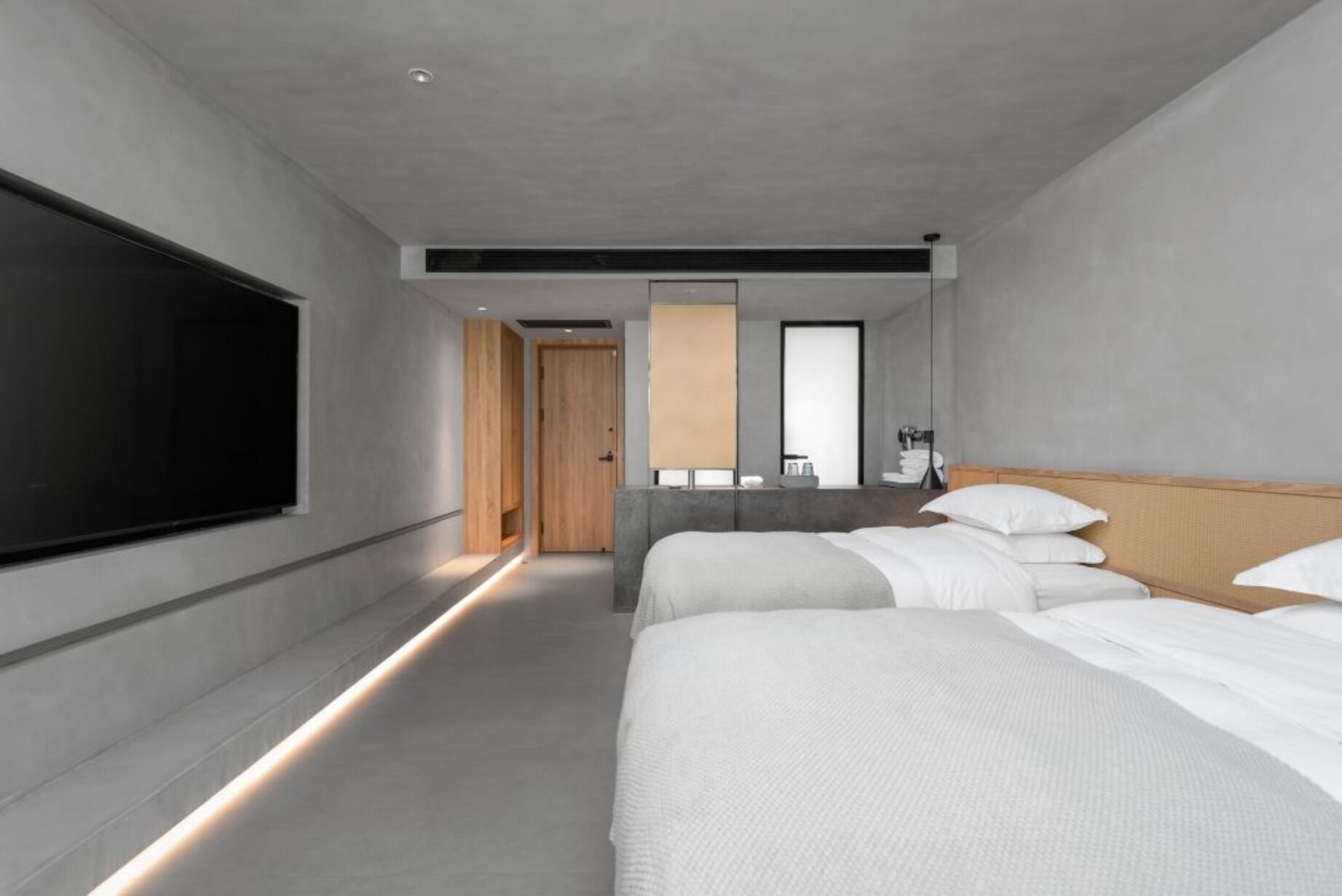 Ett modernt hotellrum med en stor platt-TV, en säng med vita sängkläder, ett träfärgat sänggavel och ett badrum med spegel och handfat.