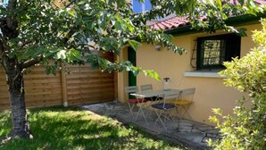 Une petite maison avec un toit en tuiles, une clôture en bois et une cour couverte d’herbe. On y trouve deux chaises pliantes et une table à l’extérieur.