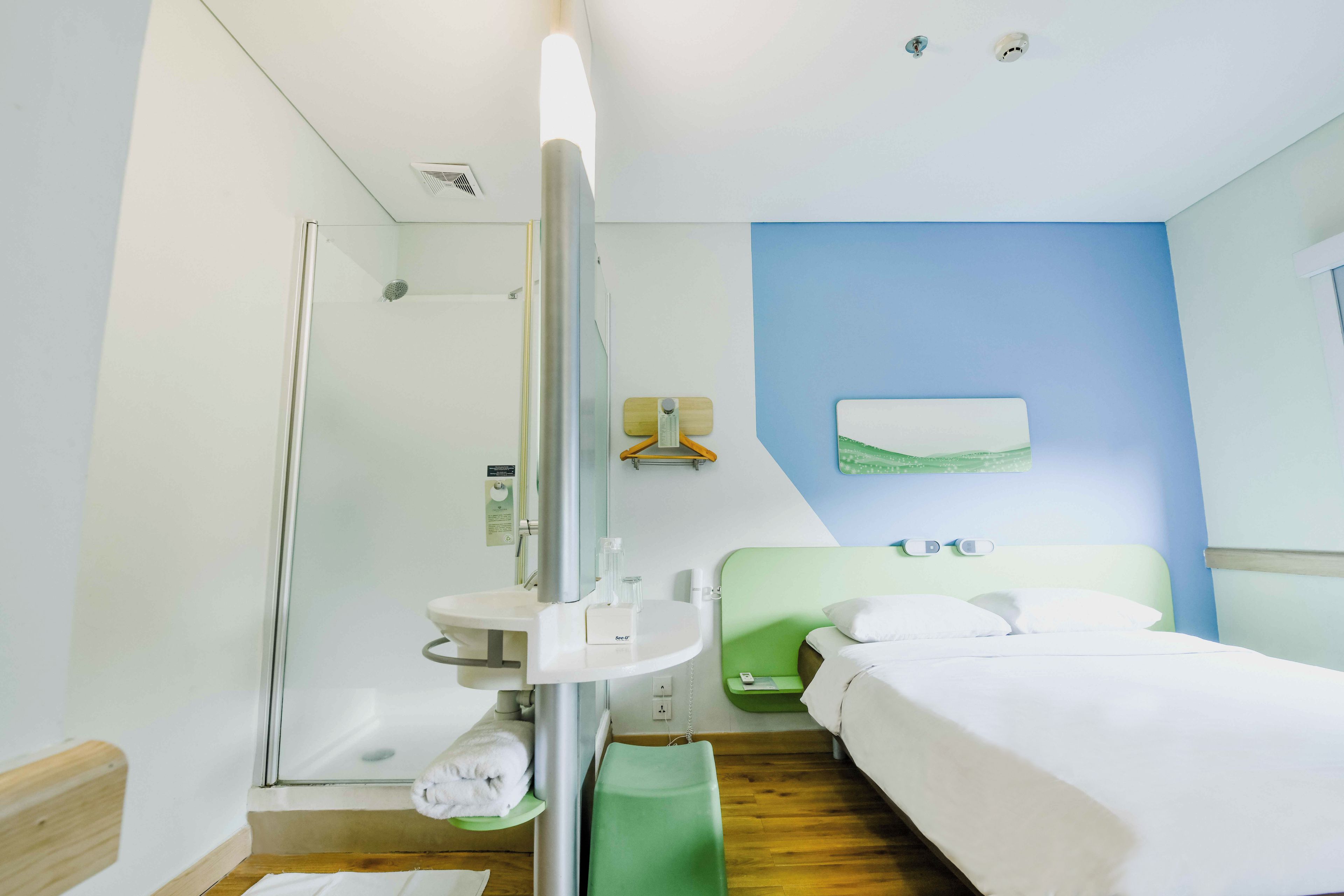 A hotel room with a shower, a bed, a bedside table, and a green bench.