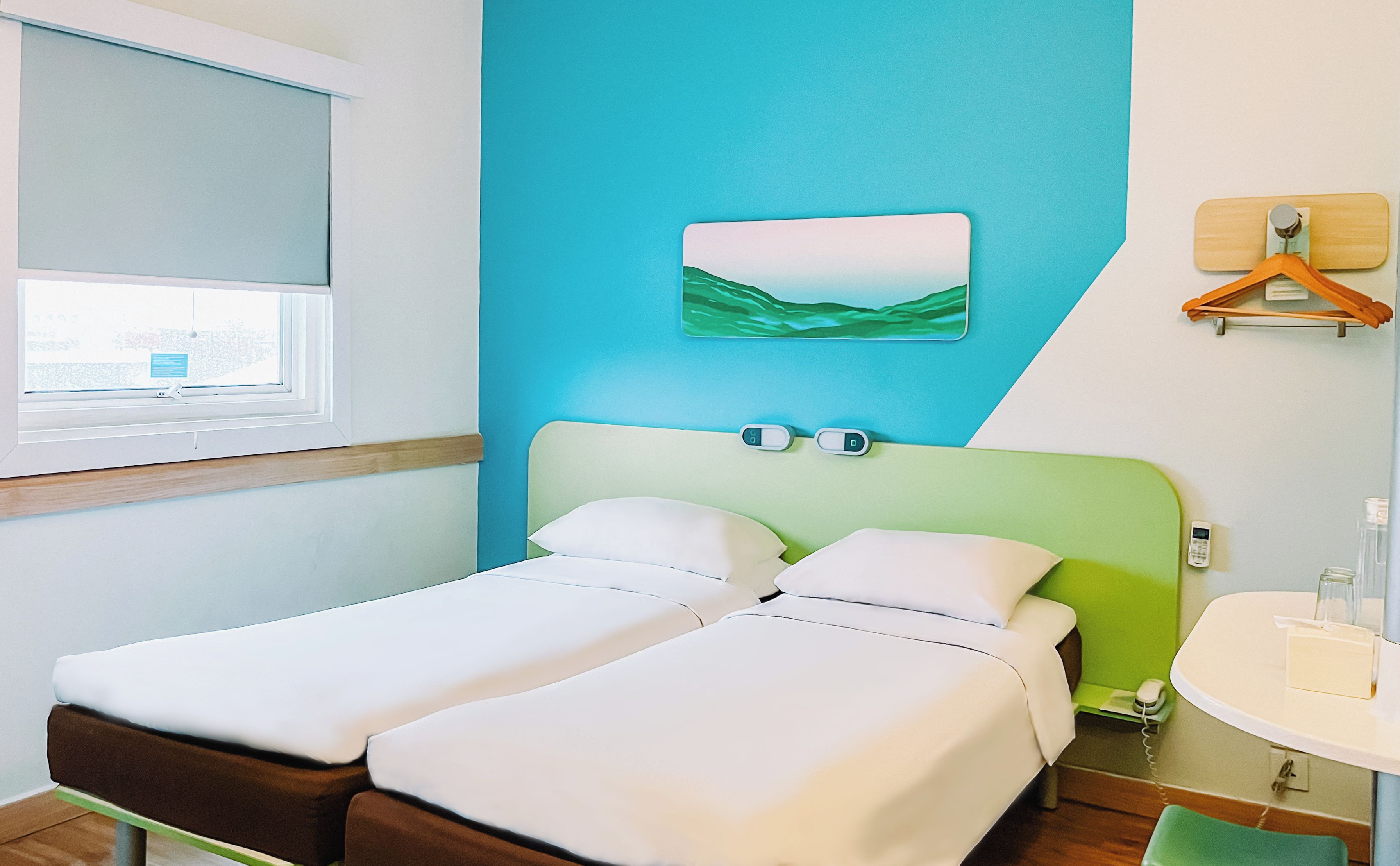 Een hotelkamer met een bed, een raam, een aan de muur hangend kunstwerk, een nachtkastje en een telefoon.