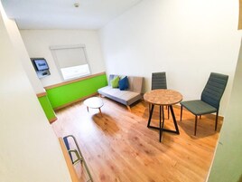 A room with a wooden floor, a round table, two chairs, a sofa with cushions, a small side table, and a window with blinds.