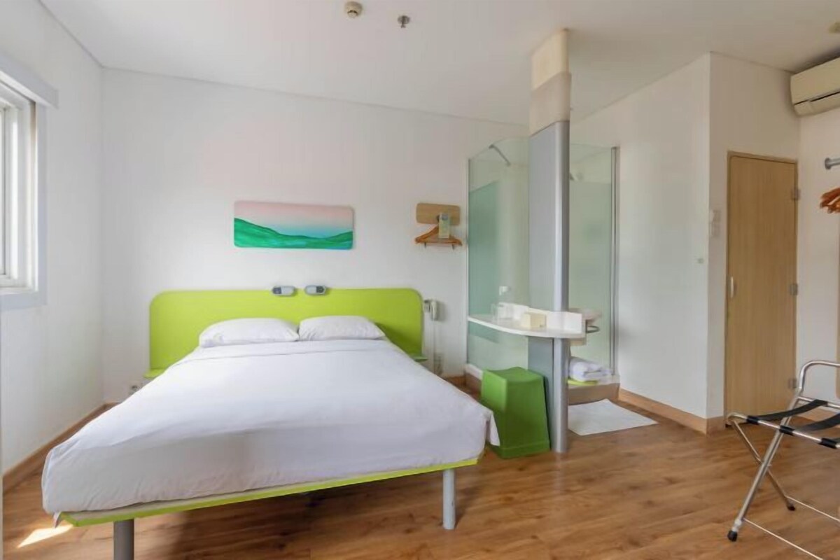 Une chambre d’hôtel moderne avec un lit doté d’une tête de lit verte, une douche en verre et un plancher en bois.
