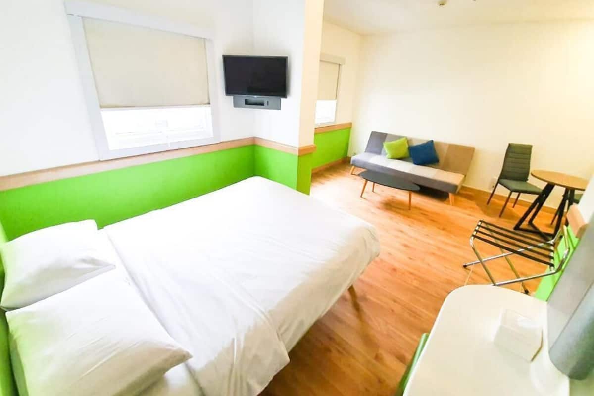 Hotelkamer met een bed, een zithoek met een bank en stoelen, een televisie en een raam met jaloezieën.
