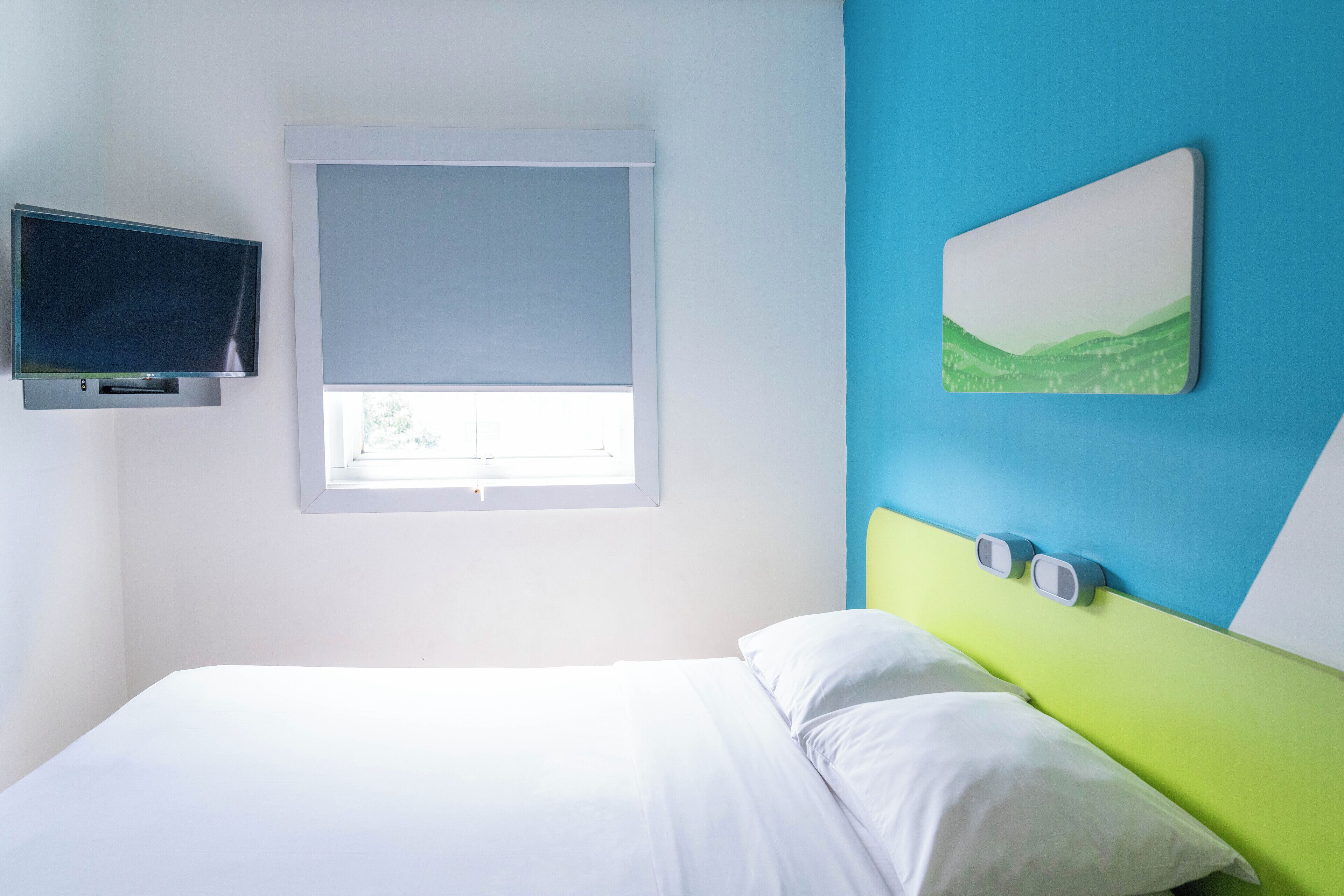 A bedroom with a bed, a television, a window with blinds, and a wall-mounted clock.