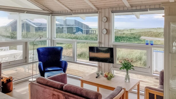 A room with large windows, a blue chair, a brown sofa, a TV, and a table with a vase and a bowl of fruit. - Stunning home in Hjørring with WiFi (Hjørring)