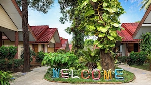 Un patio con un cartel de 'WELCOME' rodeado de abundante vegetación y un edificio con techo rojo.