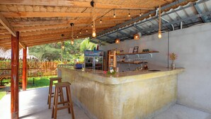 Una zona de bar de hormigón con taburetes, un refrigerador y una estantería con botellas y vasos.