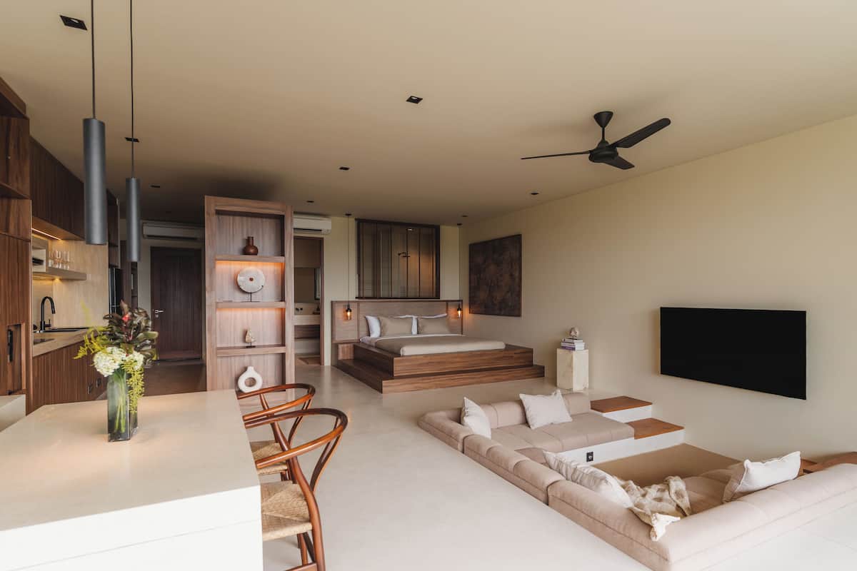 A modern living room with a sofa, a dining area with chairs, a kitchen with wooden cabinets, and a ceiling fan.