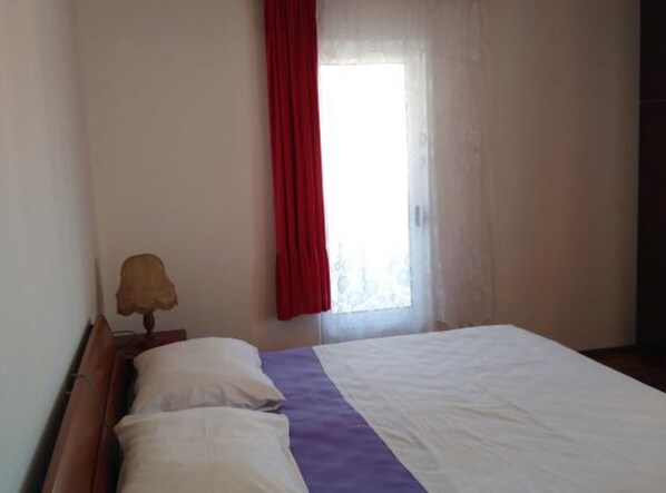 A bedroom with a bed, a nightstand, a lamp, and a window with a red curtain. - Apartmani Lucija (Okrug)