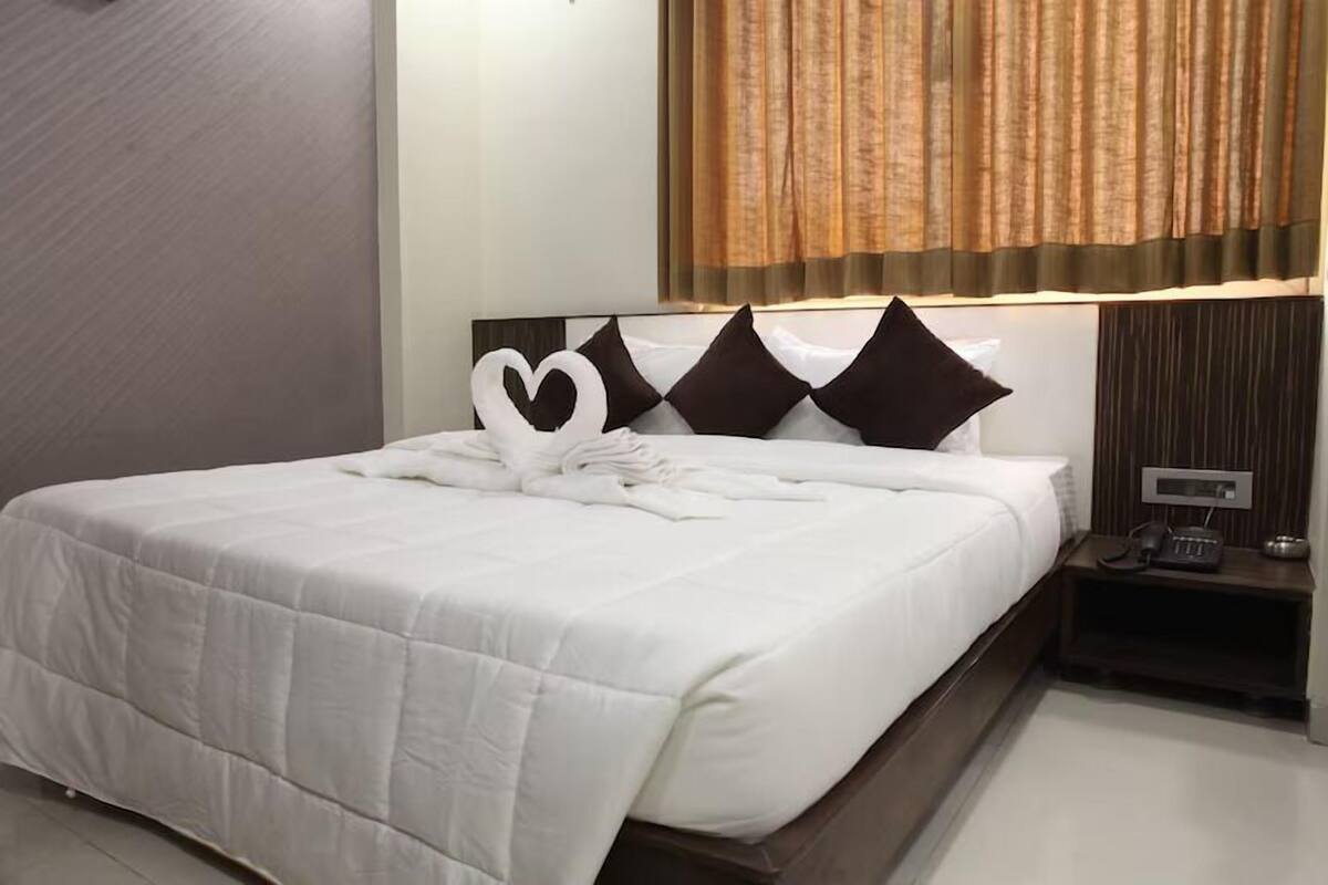 A neatly made bed with a heart-shaped towel arrangement, a bedside table with a phone, and a window with curtains.