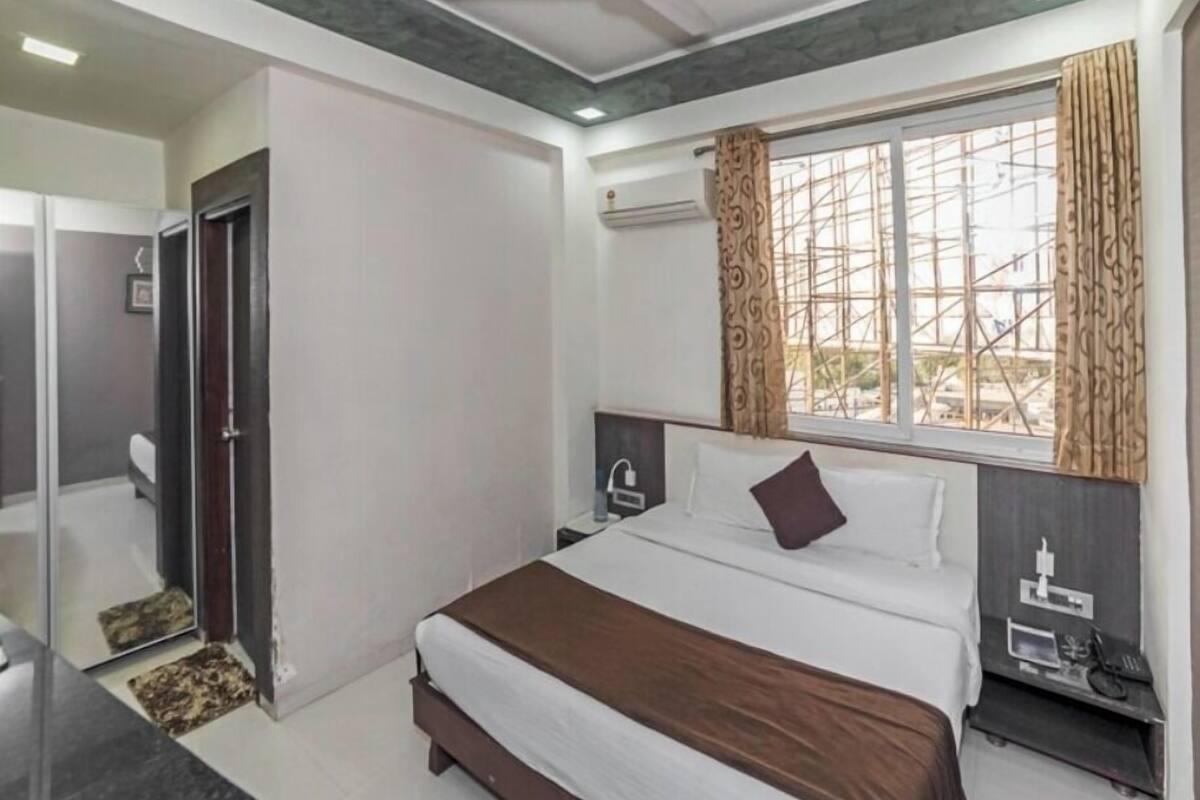 A hotel room with a bed, a bedside table, a nightstand, a window with curtains, and a door leading to a bathroom.