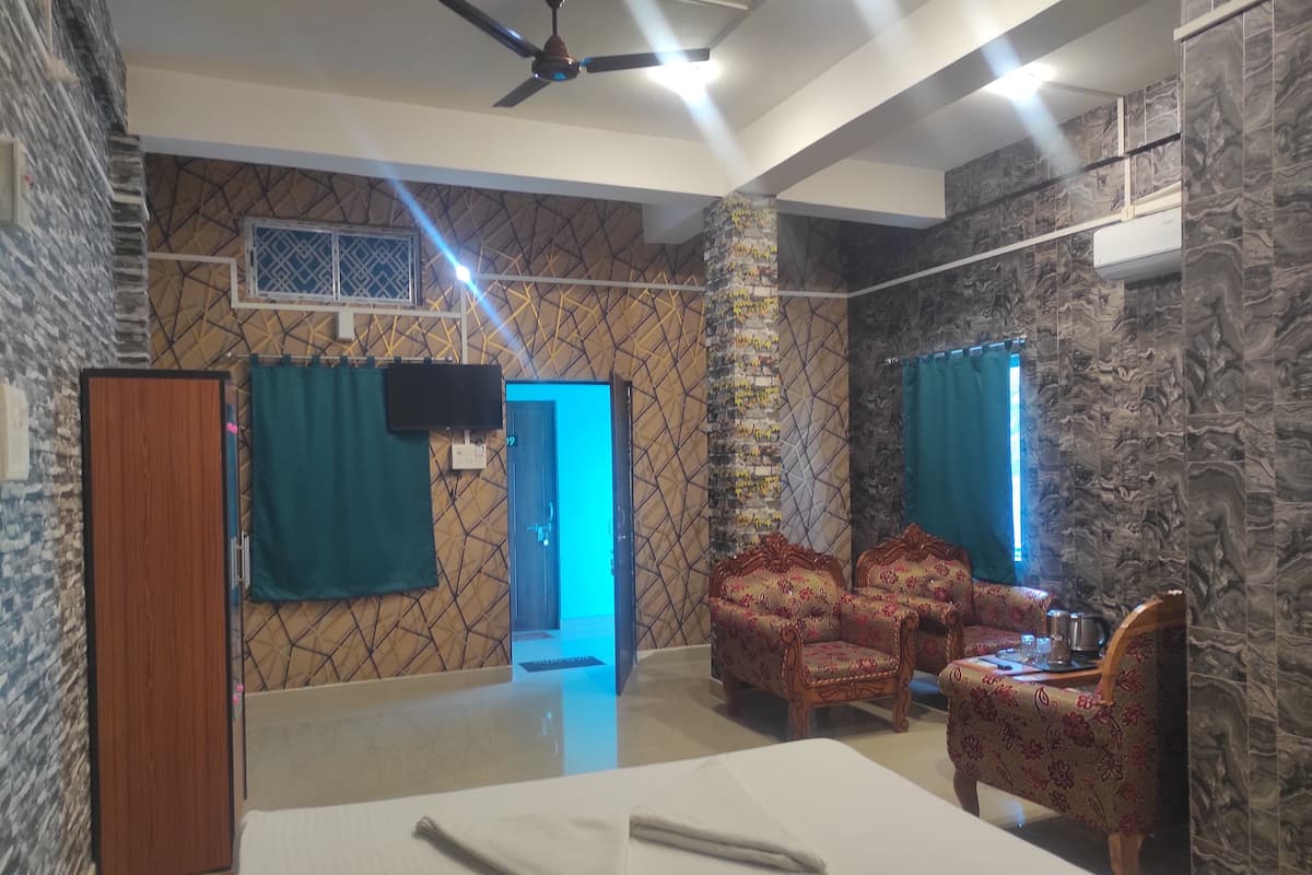 A hotel room with a bed, two armchairs, a wooden wardrobe, a television, and a ceiling fan.