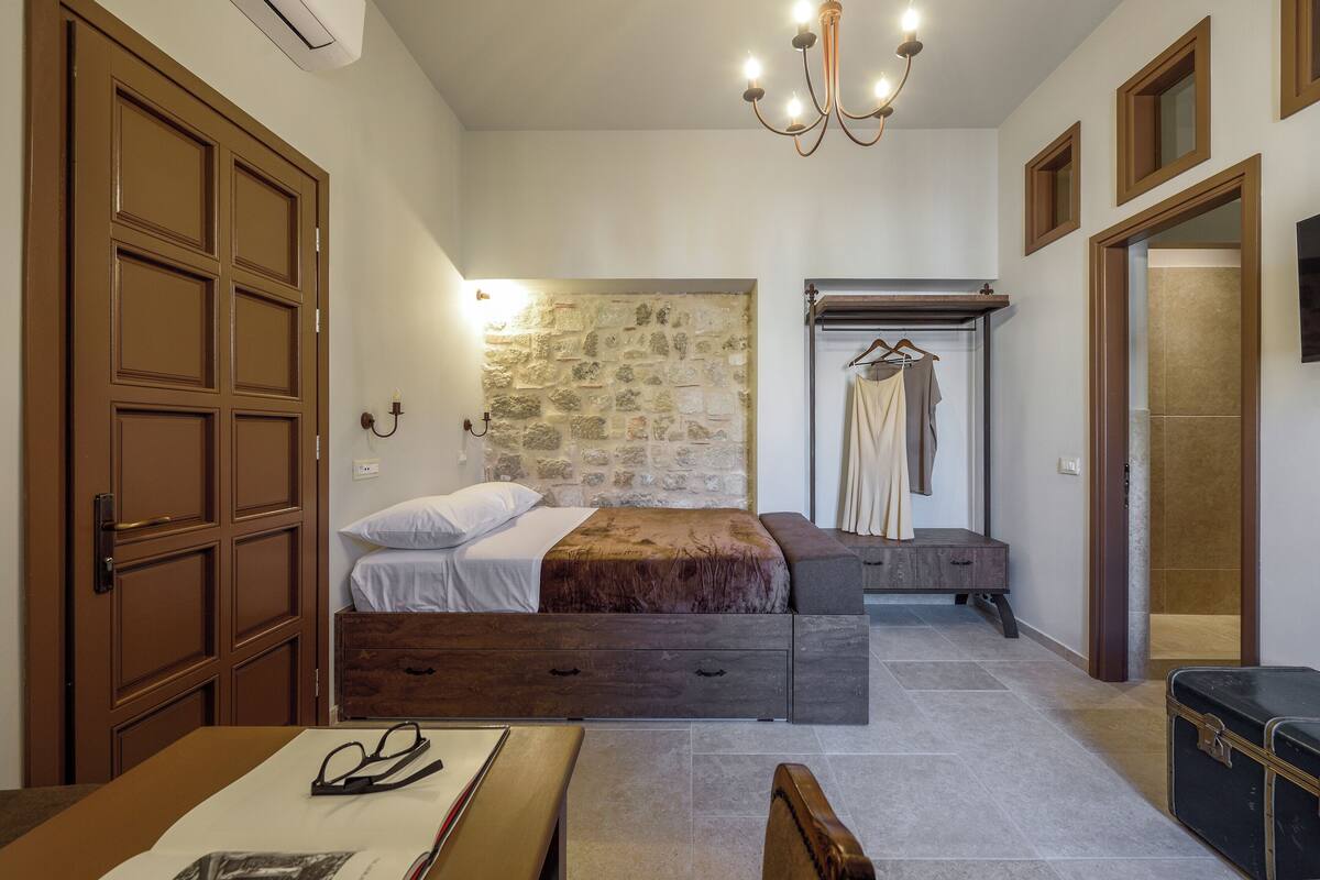 Un dormitorio con pared de piedra, cama, mesita de noche, perchero y maleta.