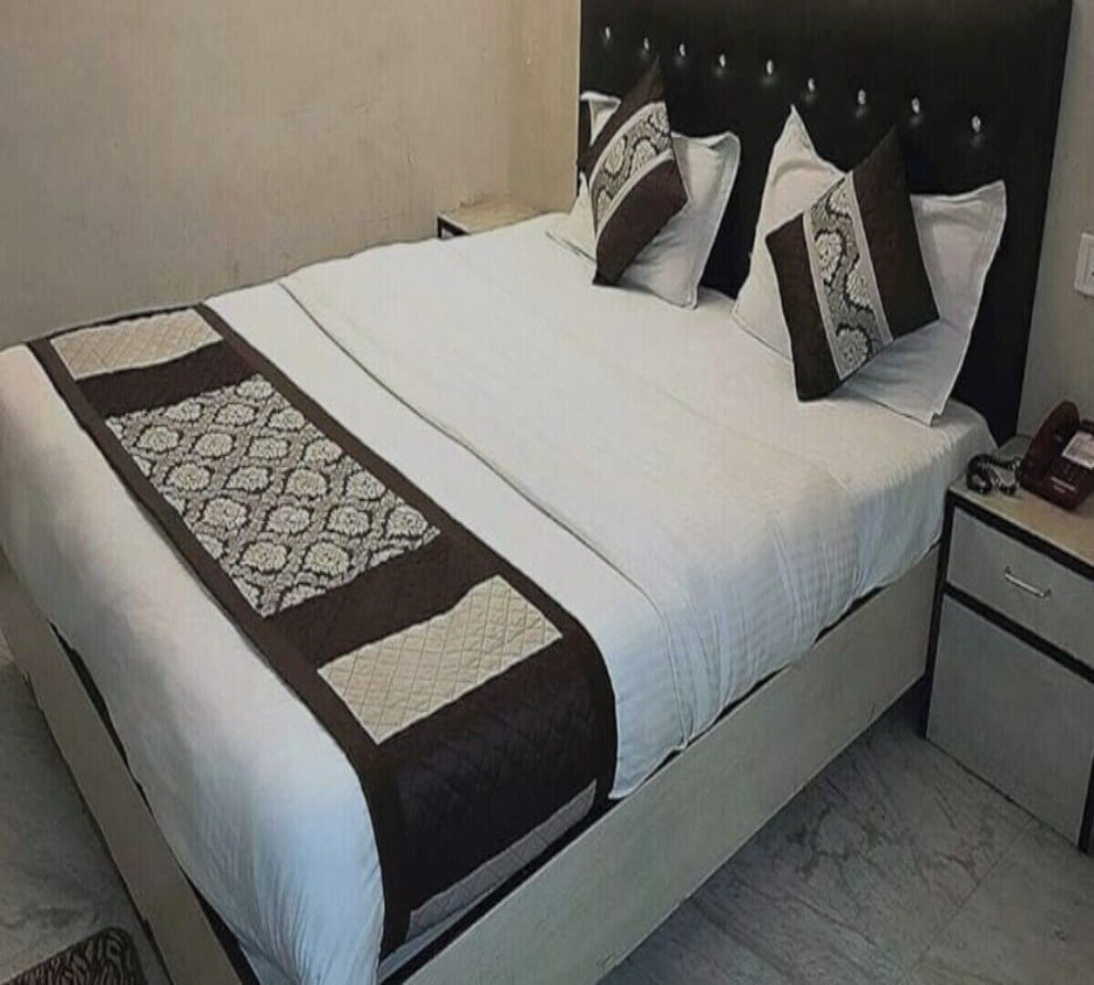 Un letto rifatto con una coperta a motivi e cuscini, un comodino con un telefono e un piccolo oggetto, e un pavimento in marmo.
