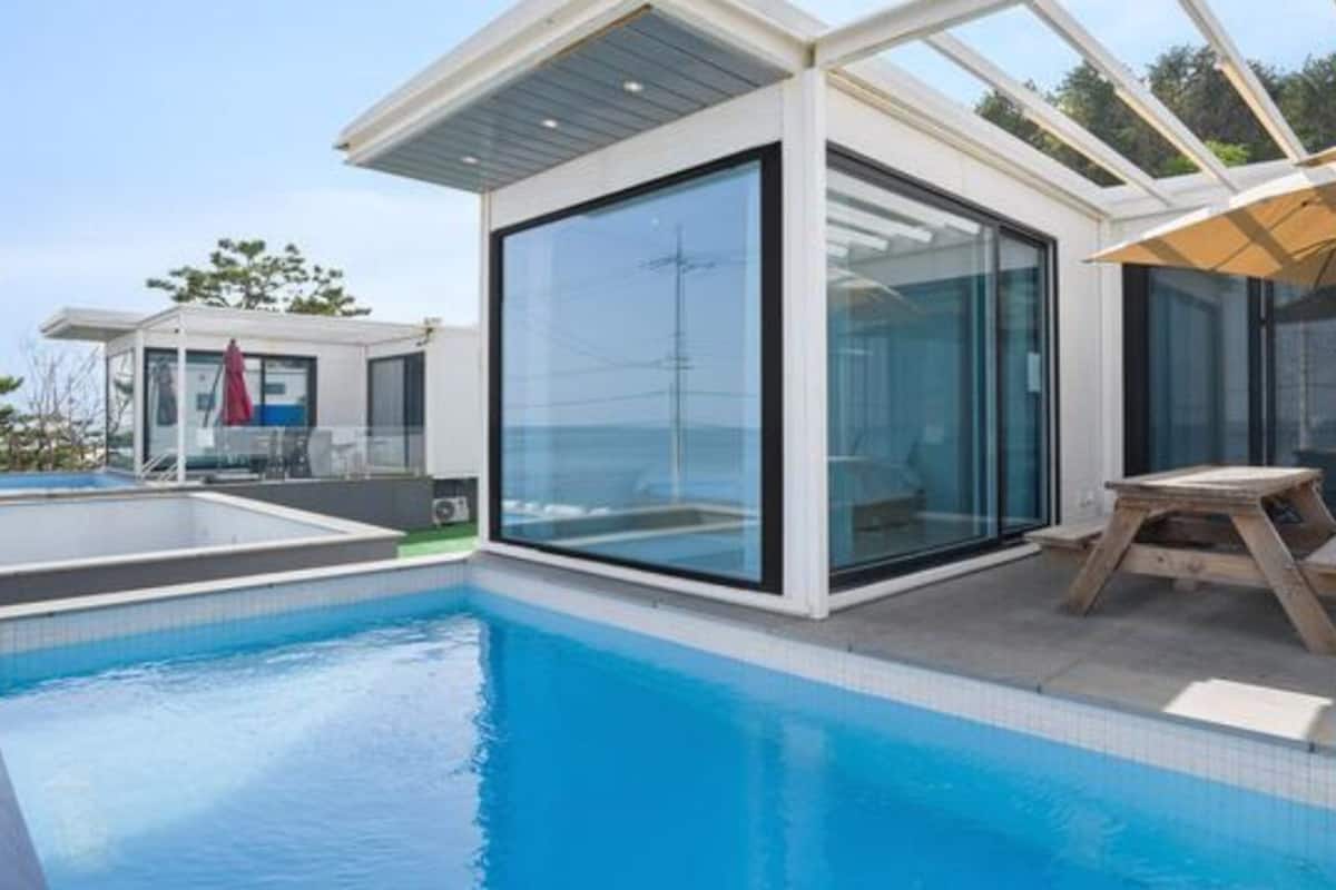Un espace piscine moderne avec des murs en verre, une table en bois et un coin salon abrité.