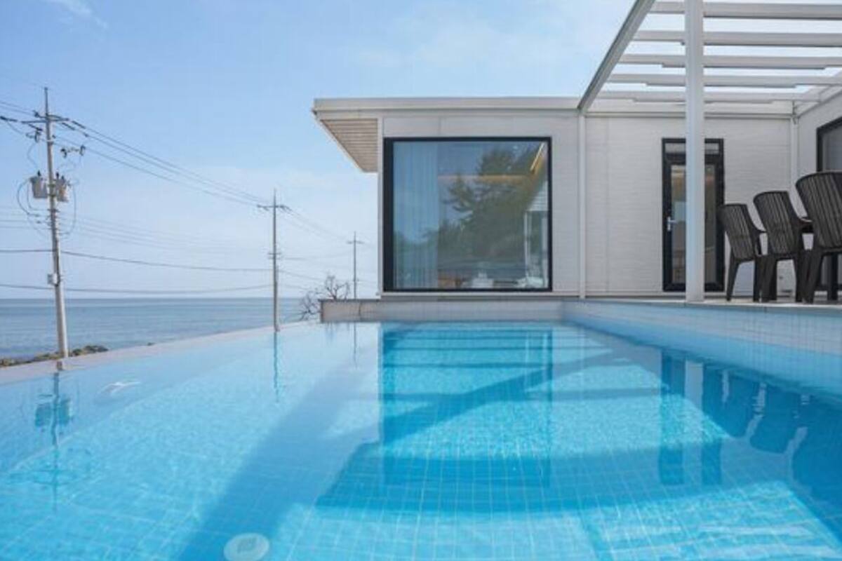 Un espace piscine moderne avec une eau d’un bleu limpide, un coin salon couvert avec des chaises, et une vue sur la mer.