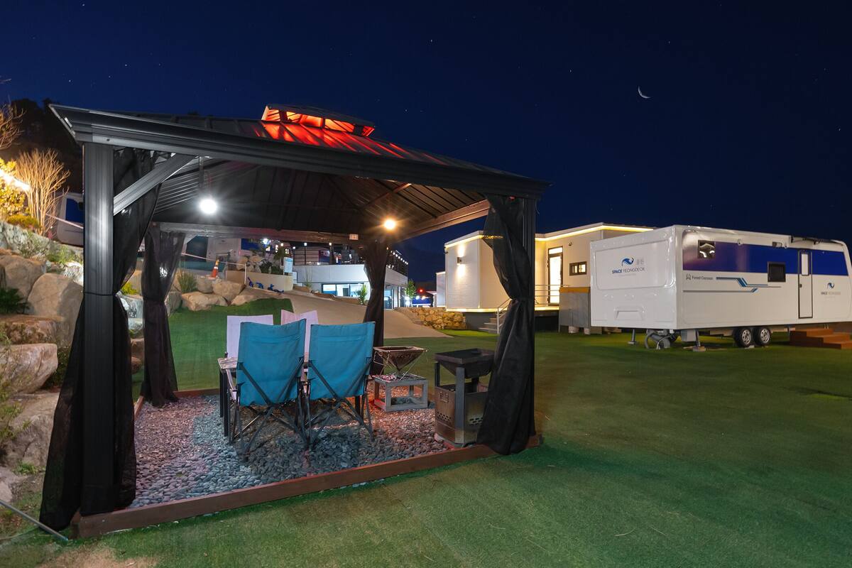 Un espace extérieur abrité avec des chaises et un barbecue, une caravane et un bâtiment dont le toit est illuminé en rouge.