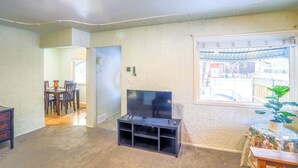 A living room with a TV on a stand, a dining area with chairs and a table, and a window with a view of the street.
