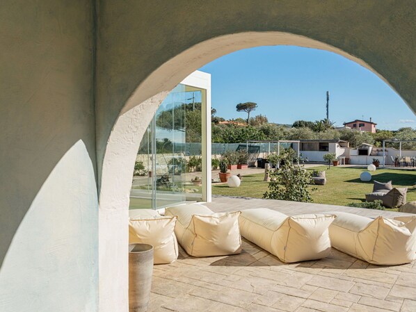 An outdoor seating area with cushions, a glass wall, and a view of a garden and buildings.