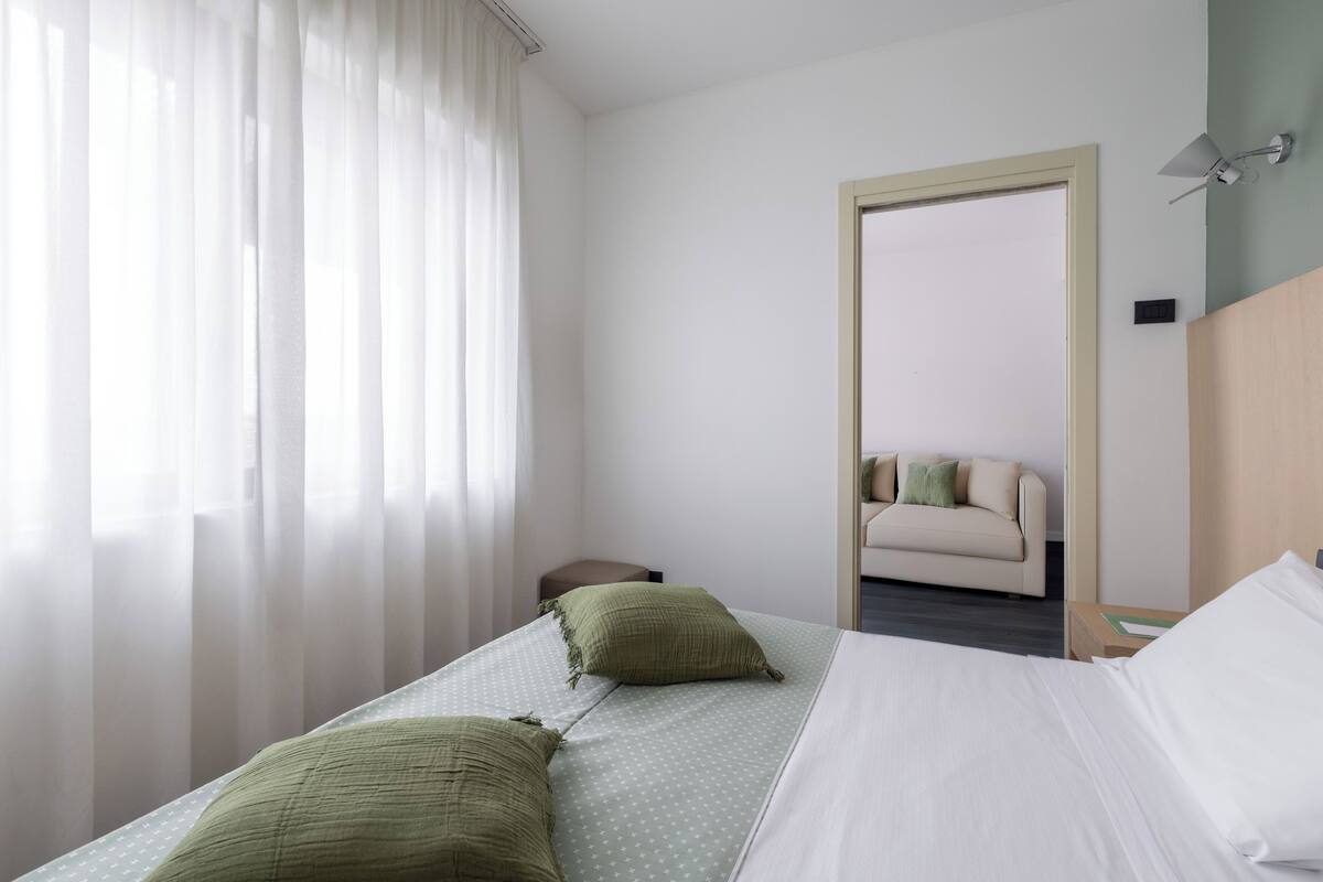 Ein Hotelzimmer mit Bett, Nachttisch, Spiegel und Fenster mit durchsichtigen Vorhängen.