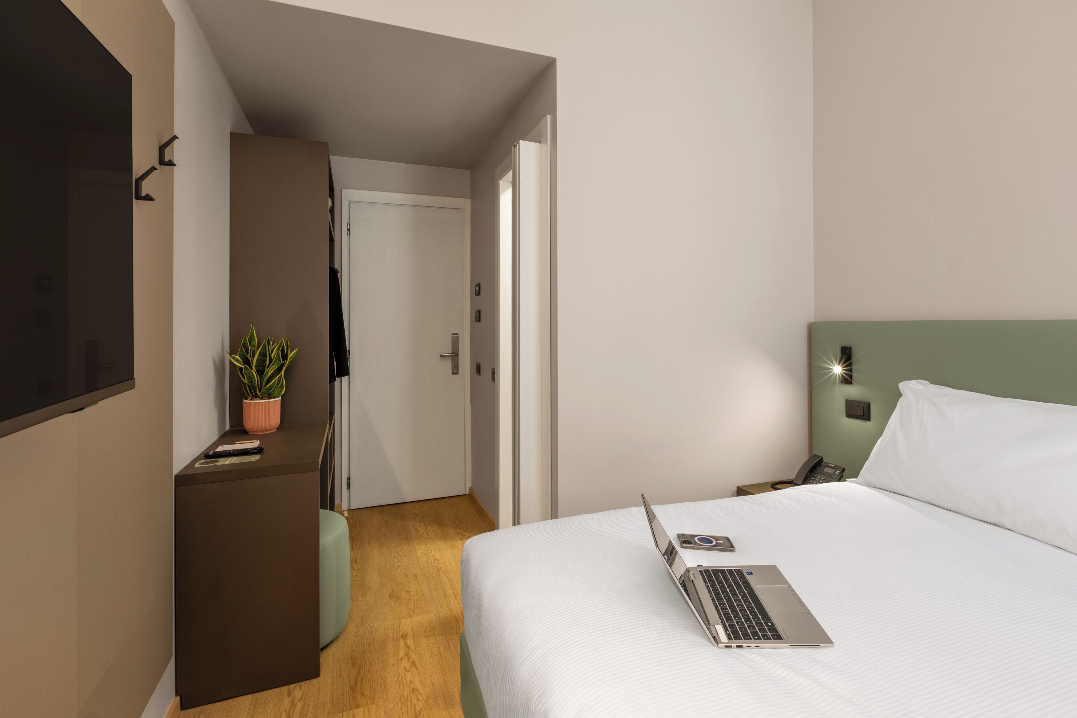 Et hotelværelse med en seng, et skrivebord med en bærbar computer, en potteplante, en telefon og en dør der fører til et andet værelse.