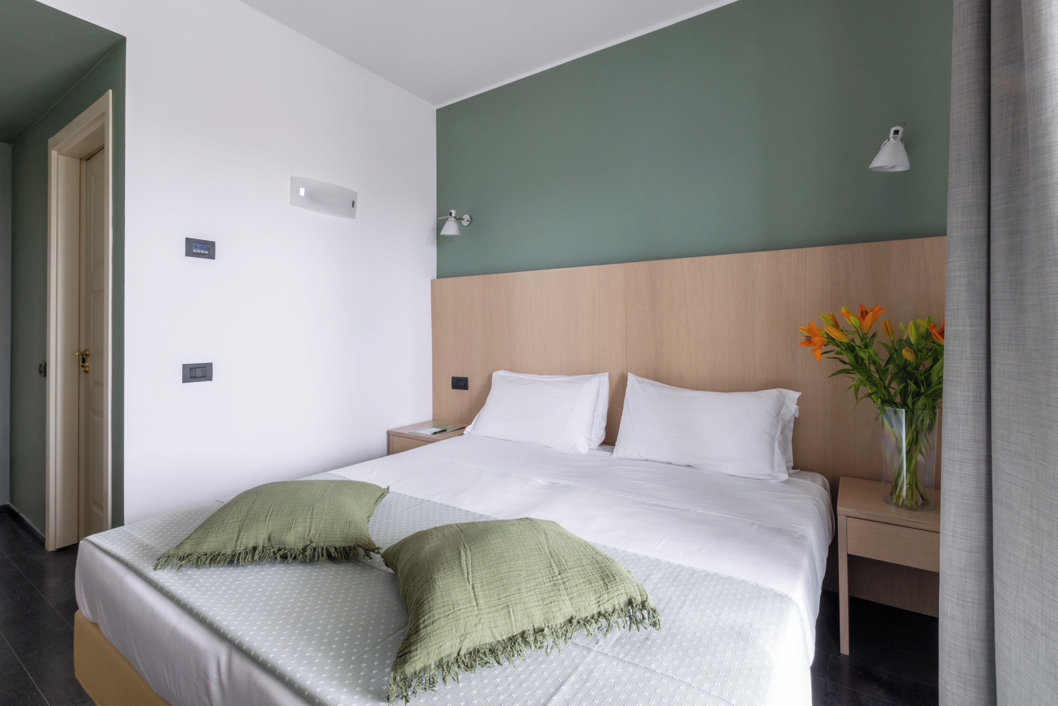 A neatly made bed with white linens and green pillows, a bedside table with a vase of flowers, and a wall-mounted lamp.