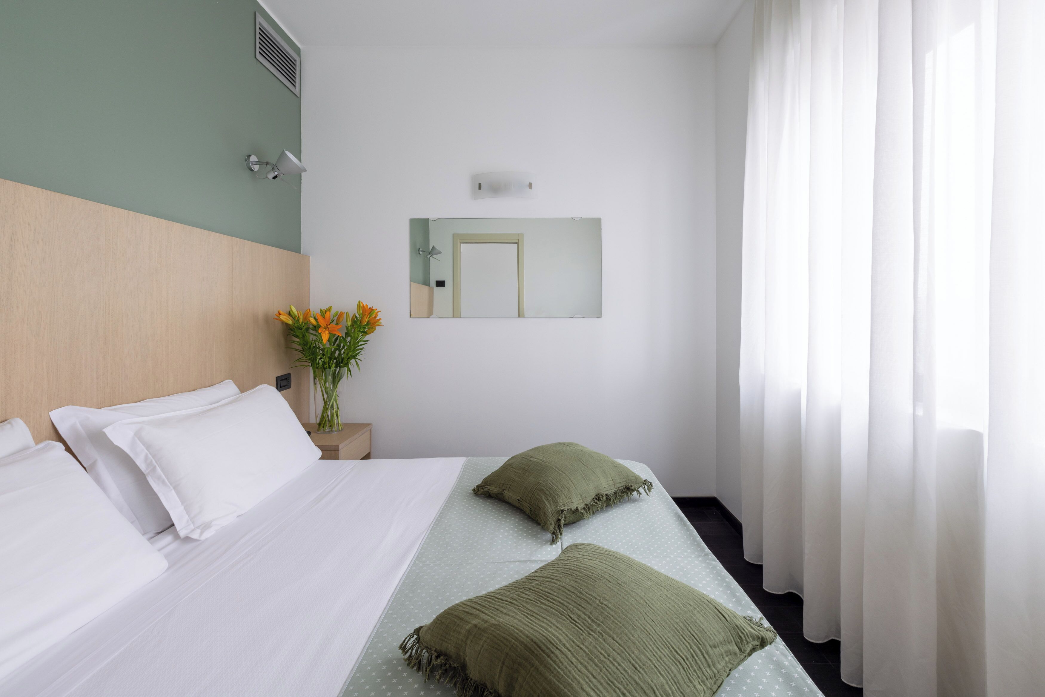 Een hotelkamer met een bed, nachtkastje, bloemenvaas, spiegel en raam met gordijnen.