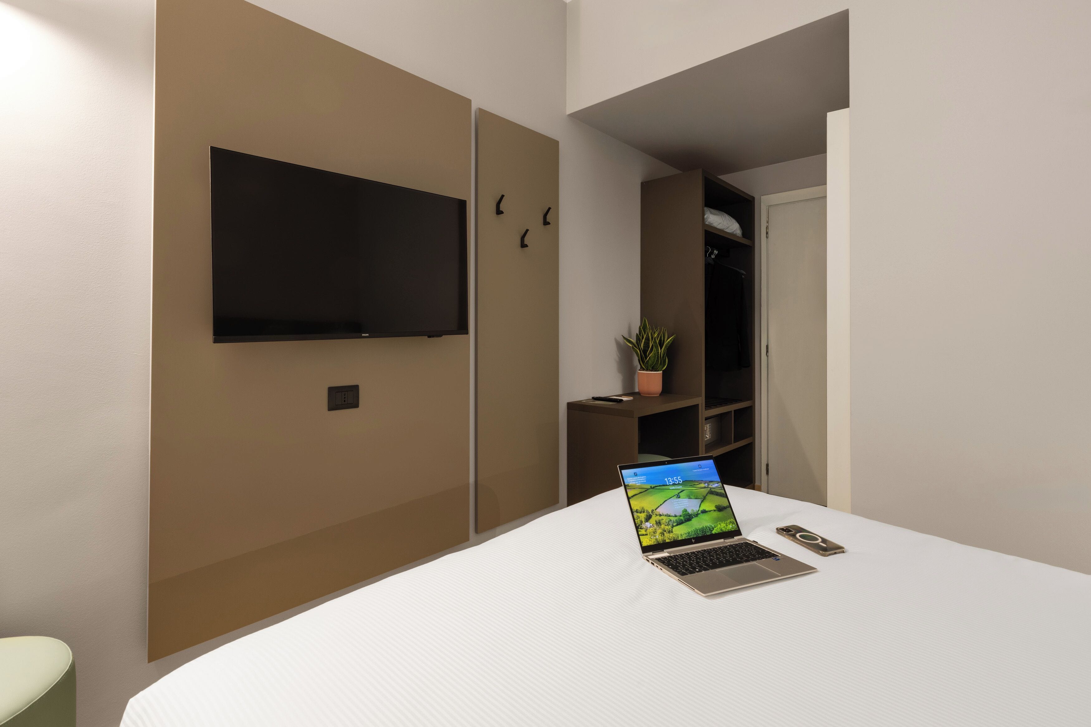 A modern hotel room with a bed, a laptop, a smartphone, a TV, and a small potted plant.