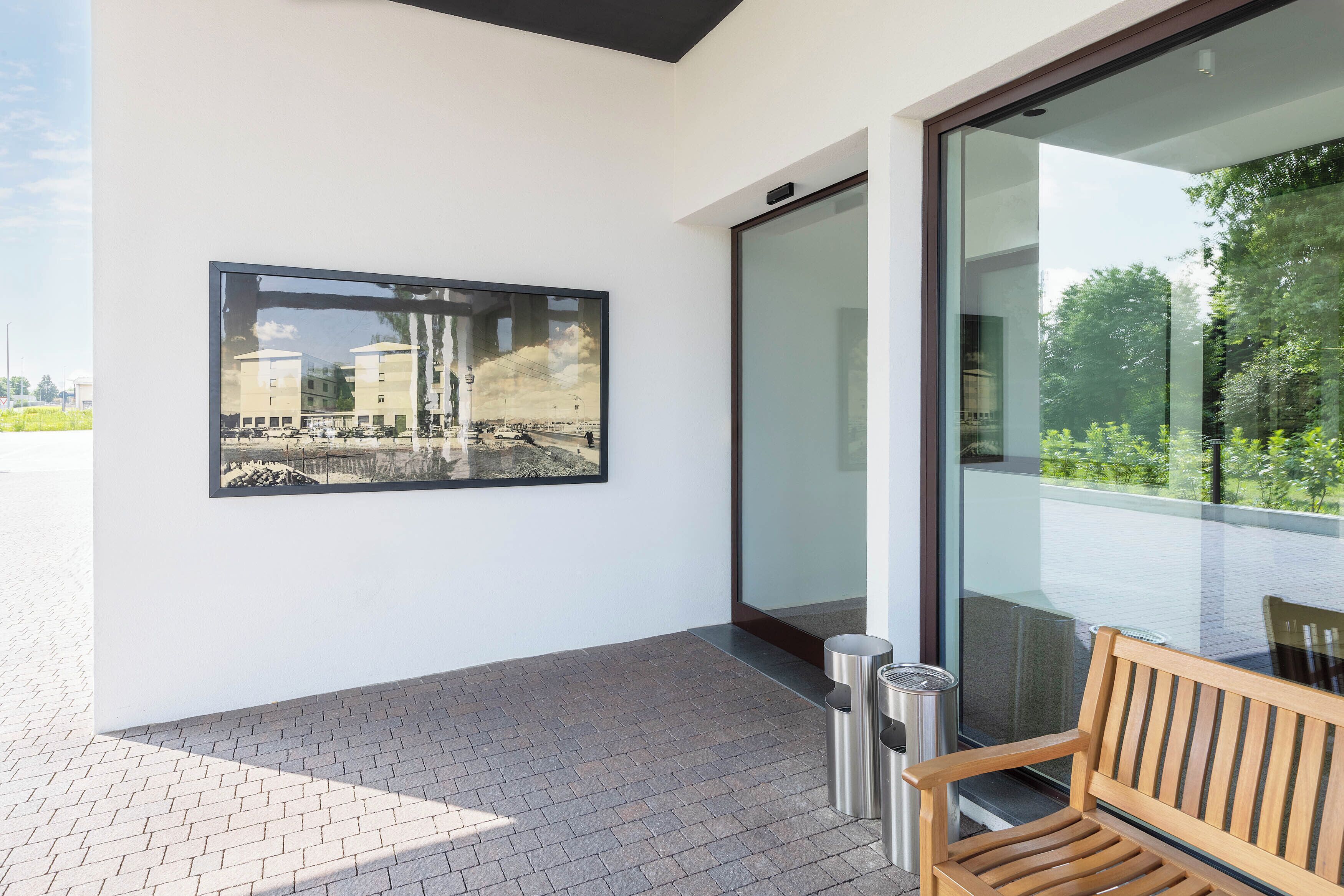 A modern building exterior with a large framed photograph on the wall, a wooden bench, and a glass door leading to a balcony.
