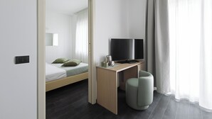 Een moderne hotelkamer met een bed, een bureau met een televisie, een koffiezetapparaat en een klein, rond poefje.