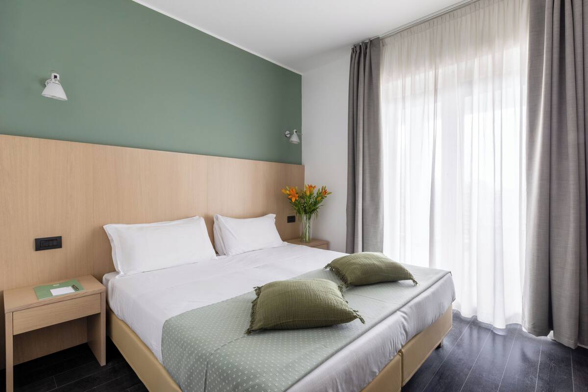 Ein Hotelzimmer mit Bett, Nachttisch, Lampe und einem Fenster mit Vorhängen.